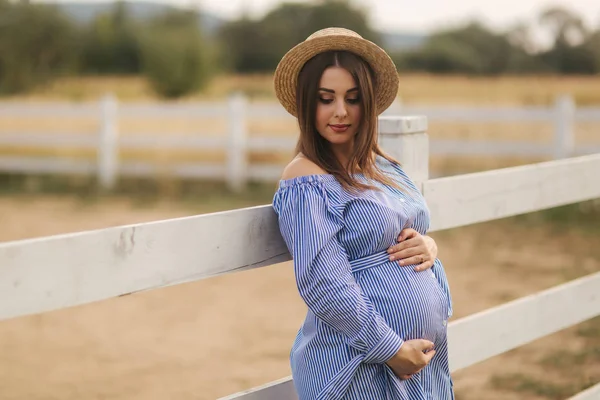 Bella donna incinta in abito blu e cappello a maglia stand vicino alla recinzione nella fattoria. Si mise le mani sulla pancia e sorrise — Foto Stock