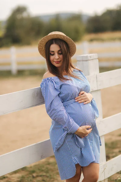 Красивая беременная женщина в синем платье и вязаной шляпе стоять рядом с модой на ферме. Она положила руки на живот и улыбнулась. — стоковое фото