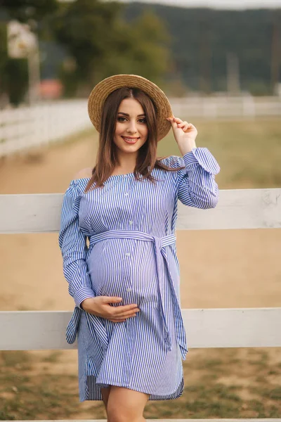 Belle femme enceinte en robe bleue et chapeau tricoté debout près de la clôture dans la ferme. Elle a mis les mains sur son ventre et sur son chapeau — Photo