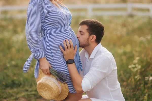 Ein Mann küsst Bauch einer schwangeren Frau in der Natur. Mann im weißen Hemd und Frau im blauen Kleid — Stockfoto