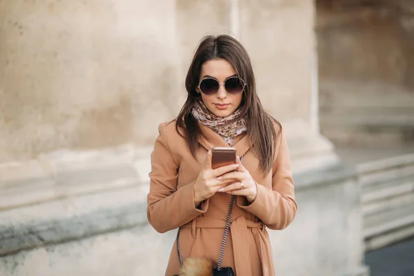 Stijlvolle meisje in een bruine jas en een sjaal met een bril. Met behulp van een telefoon. Make-up — Stockfoto