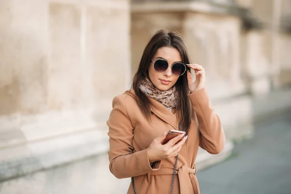 Stylowa dziewczyna w brązowy płaszcz, szalik z okularami. Przy użyciu telefonu. Makijaż — Zdjęcie stockowe