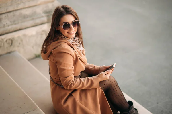 Стильная девушка в коричневом пальто и шарф в очках. С помощью телефона. Макияж — стоковое фото