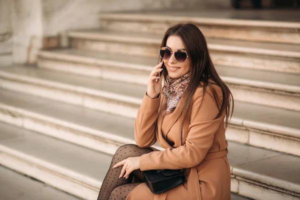 Stylish girl in a brown coat and a scarf with glasses. Using a phone. Makeup