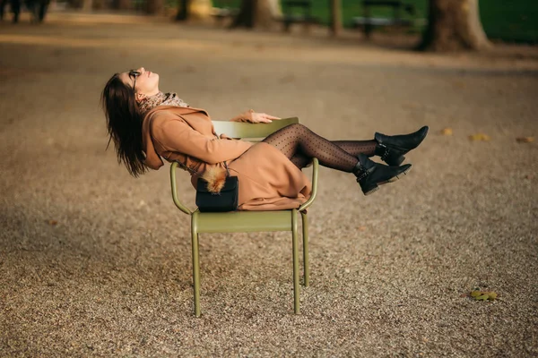 A young girl in a brown coat sits in the middle of the park on a khaki stool. Girl relax and have good day — Stock Photo, Image