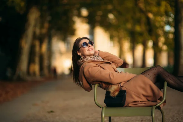Una joven con un abrigo marrón se sienta en medio del parque en un taburete caqui. Chica relajarse y tener un buen día —  Fotos de Stock