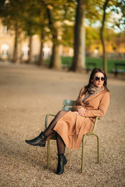 Молодая девушка в коричневом пальто сидит посреди парка на стуле хаки — стоковое фото