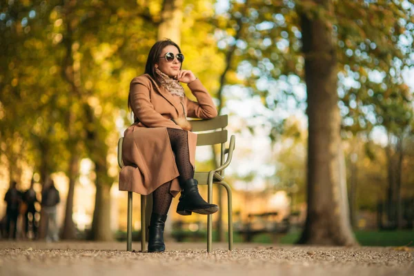 Młoda dziewczyna w brązowy płaszcz siedzi w środku parku na stołku khaki — Zdjęcie stockowe