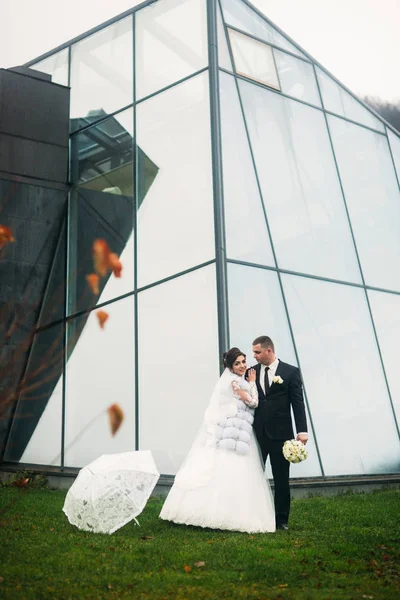 Groom and bride near the mirror building in in bad weather. They use umbrella