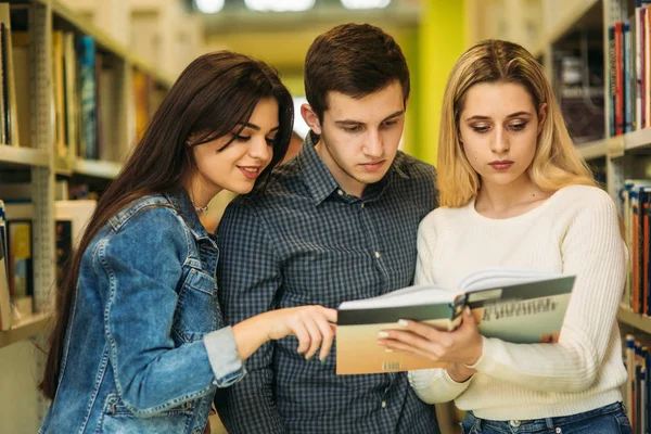 Grupo de estudiantes quieren encontrar un poco de literatura útil para la preparación para el examen universitario. Dos chicas y un chico revisan el libro. — Foto de Stock