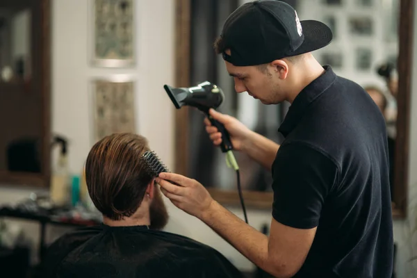 Master cuts hair and beard of men in the barbershop and uses a hair dryer