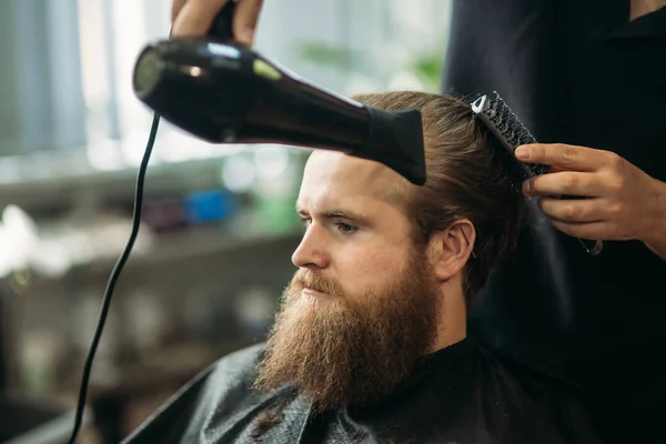 Master cuts hair and beard of men in the barbershop and uses a hair dryer.