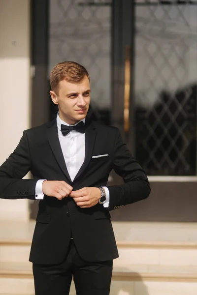 Fashion groom in trendy suit posing to photographer. Blond hair man with bow tie and other accsessories stand by the beautiful hotel — Stock Photo, Image