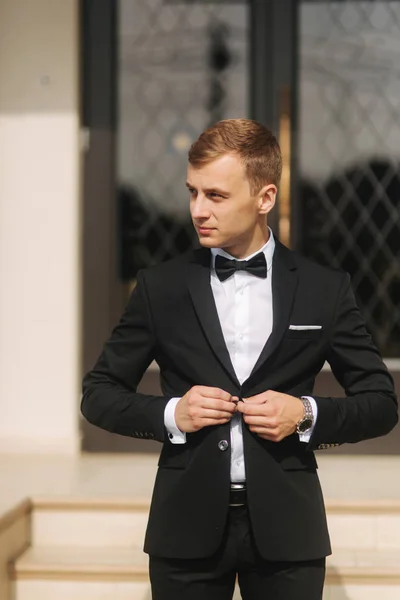 Fashion groom in trendy suit posing to photographer. Blond hair man with bow tie and other accsessories stand by the beautiful hotel — Stock Photo, Image