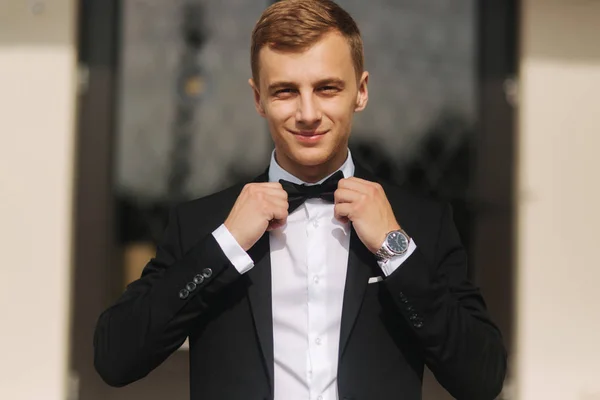 Novio de moda en traje de moda posando para el fotógrafo. Hombre de pelo rubio con corbata de lazo y otros accesoriosstand junto al hermoso hotel —  Fotos de Stock