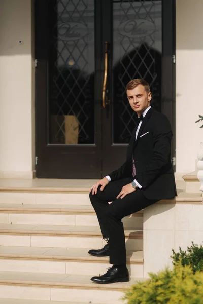 Fashion groom in trendy suit posing to photographer. Blond hair man with bow tie and other accsessories stand by the beautiful hotel — Stock Photo, Image