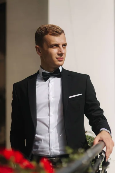 Fashion groom in trendy suit posing to photographer. Blond hair man with bow tie and other accsessories stand by the beautiful hotel — Stock Photo, Image