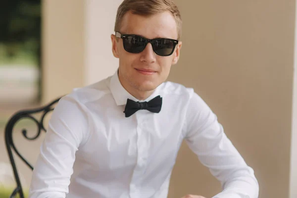 Hombre guapo con camisa y pajarita usa el teléfono. el hombre rubio del pelo afuera. Hombre sist por la mesa y probar en gafas de sol —  Fotos de Stock