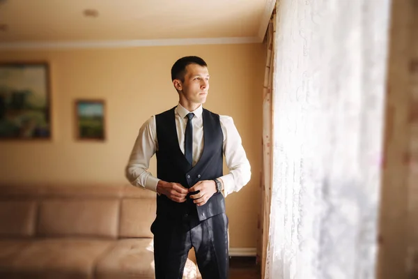 Handsome groom in suit stand near the window. He wears a tie and watch — Stock Photo, Image