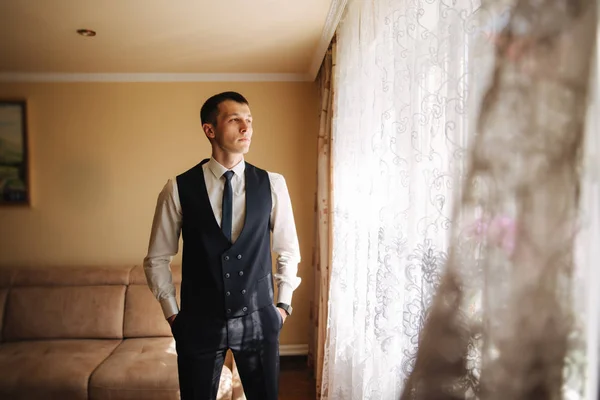 Handsome groom in suit stand near the window. He wears a tie and watch — Stock Photo, Image