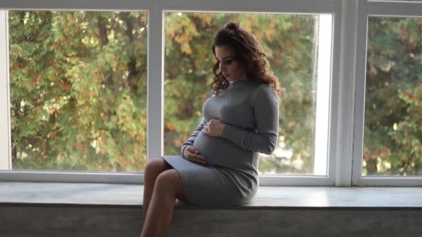 A pregnant woman sitting on the windowsill near the window at home. She dressed in beautiful maternity dress — Stock Video