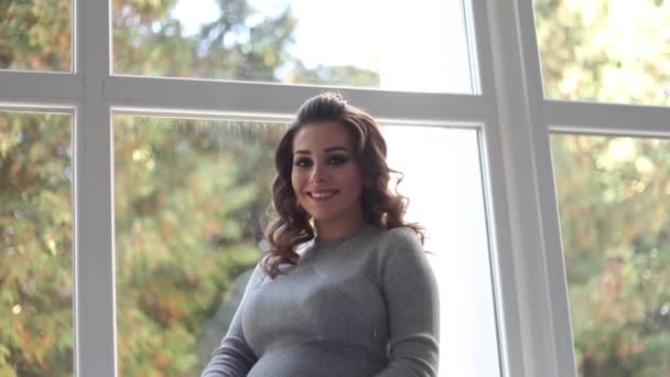 임신한 여 자가 집에서 창문 가까이 창턱에 앉아. 그녀는 아름 다운 출산 드레스 입고 — 비디오