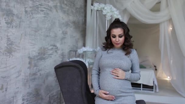 Schöne schwangere Frau auf dem Sessel sitzen und legte ihre Hände auf ihren Bauch. Frau in elegantem Kleid. schöne Frisur und Make-up — Stockvideo