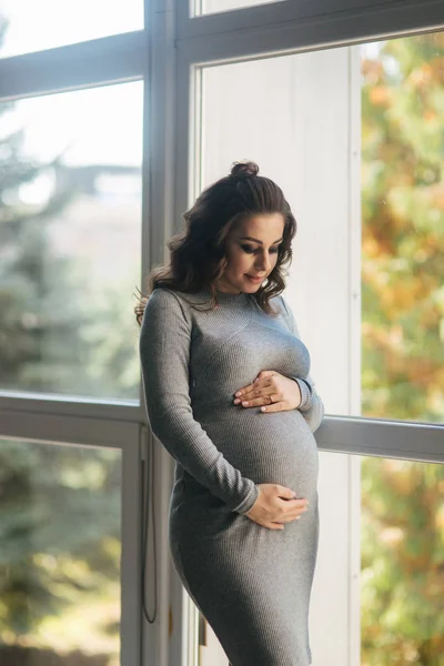 Mujer embarazada en un hermoso vestido gris de pie cerca de la ventana y acariciando su barriga —  Fotos de Stock