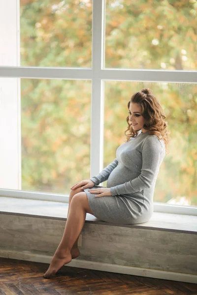 Eine schwangere Frau, die zu Hause auf der Fensterbank neben dem Fenster sitzt. sie trug ein wunderschönes Umstandskleid — Stockfoto