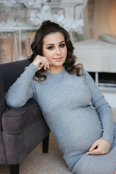 Gravid kvinna sitta nära fåtöljen. Vacker kvinna väntar en baby. Elegant gravid modell hemma — Stockfoto