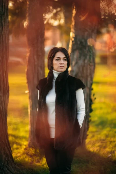Dark haired woman stand in front of tree. Woman has bob haircut.