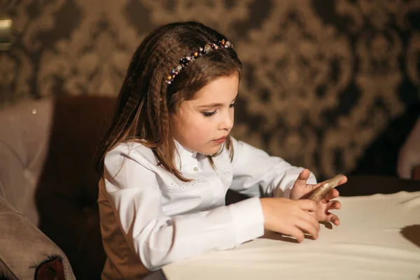 Mała dziewczynka używać telefonu w kawiarni. Składać się ona na krześle przy stole — Zdjęcie stockowe