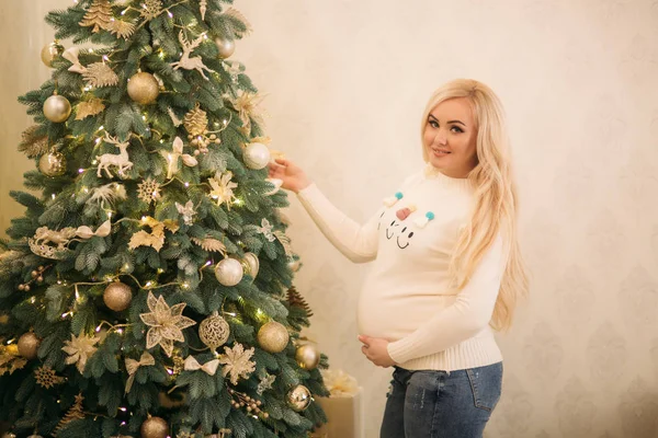 Całkiem w ciąży Blondynka w Christmas sweter stoi w pobliżu choinki — Zdjęcie stockowe