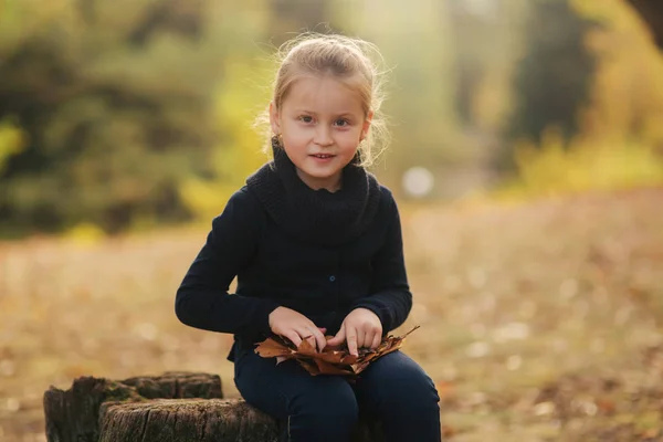 Una bambina in abiti neri è seduta su un ceppo e gioca con le foglie autunnali. Ragazza capelli biondi — Foto Stock