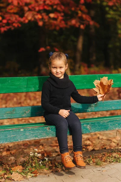 La niñita se sienta en el banco y mira las hojas. Recoge hojas y las lleva con ella. Bosque de otoño — Foto de Stock
