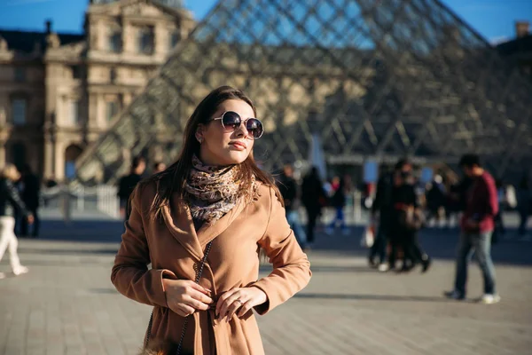 Uma jovem de casaco castanho e cachecol fica no fundo da cidade de Paris. Outono é tempo ensolarado, turistas — Fotografia de Stock