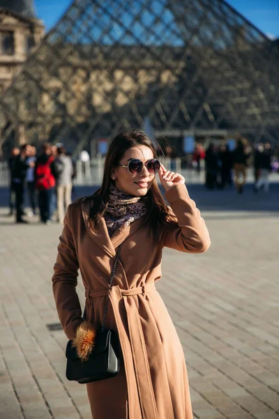 Молодая девушка в коричневом пальто и шарфе стоит на фоне города Парижа. Осень - солнечная погода, туристы — стоковое фото