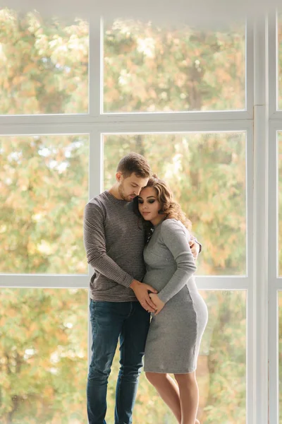 男人拥抱怀孕的妻子, 把手放在她的肚子上。他们站在窗户附近 — 图库照片