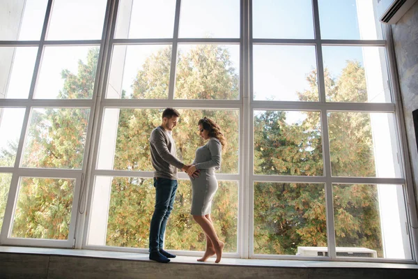 Schönes Paar steht vor großem Fenster im Atelier. Schwangere mit ihrem Mann — Stockfoto