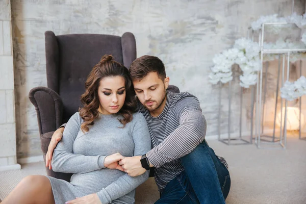 Coppia felice siede vicino alla sedia nella loro casa. Donna incinta con il suo amato marito. nuovo anno — Foto Stock