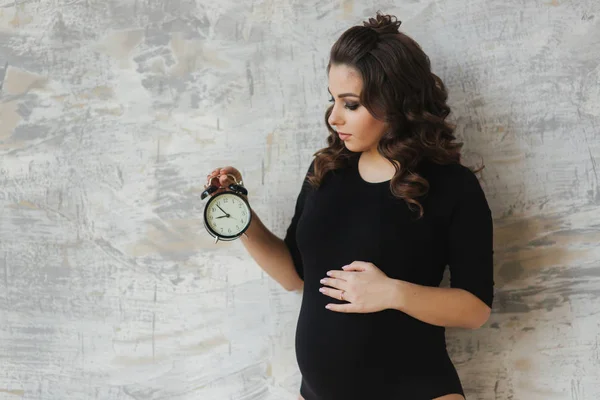 Ritratto di bella donna incinta in body nero. Tiene l'orologio con le mani simboliche alludendo al tempo rimanente befour il bambino in nato. Donna guarda l'orologio — Foto Stock