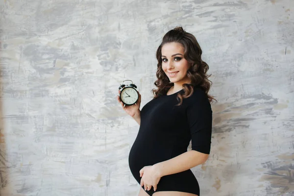 युवा गर्भवती महिला अपने हाथों में एक घड़ी रखती है। भविष्य की मां का सुंदर चित्र। महिला एक बच्चे, बच्चे की प्रतीक्षा कर रही है — स्टॉक फ़ोटो, इमेज