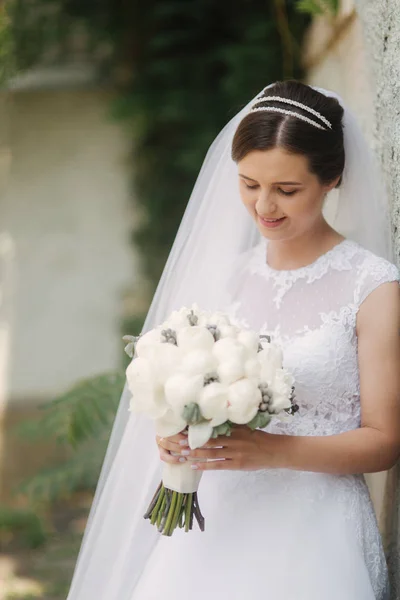 Junge Braut in schönem weißen Hochzeitskleid stehen in der Nähe der Mauer. Sie hält einen Blumenstrauß in den Händen — Stockfoto