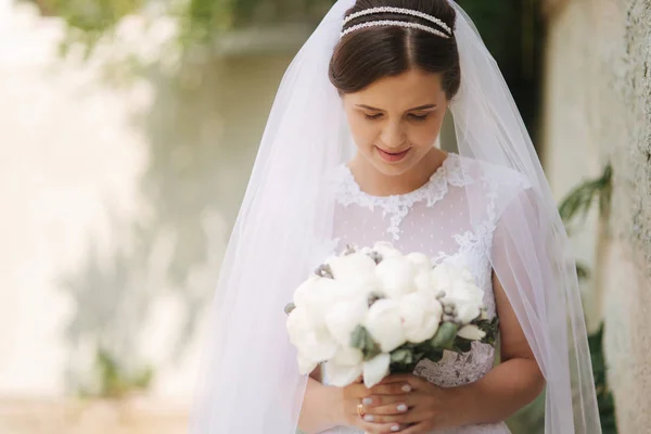 Giovane sposa in bellissimo abito da sposa bianco stand vicino al muro. Tiene un mazzo di fiori in mano — Foto Stock