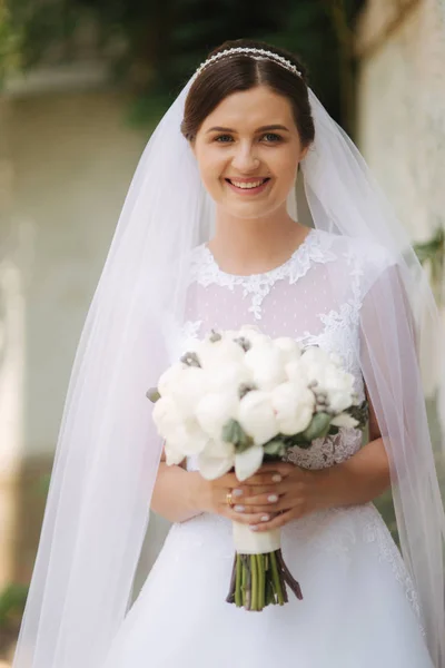 Jovem noiva em belo vestido de noiva branco ficar perto da parede. Ela segura um buquê de flores nas mãos — Fotografia de Stock