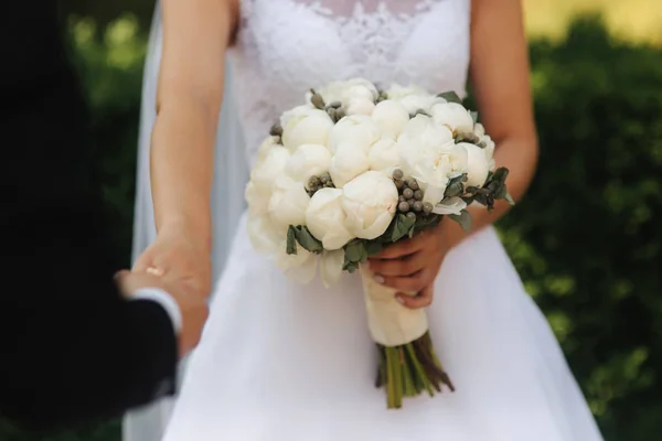 Giovane sposa in bellissimo abito da sposa bianco stand vicino al muro. Tiene un mazzo di fiori in mano — Foto Stock