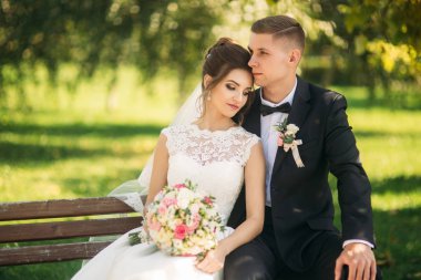 Düğün giysileri içinde genç bir çift bir parkta bir bankta oturuyor. Güneşli havalarda