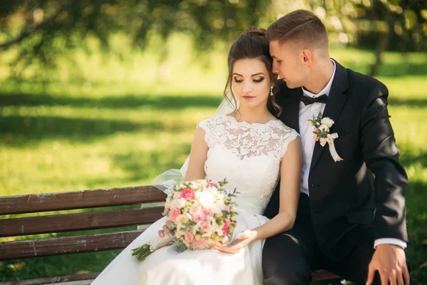 Una giovane coppia in abiti da sposa è seduta su una panchina in un parco. Tempo soleggiato — Foto Stock