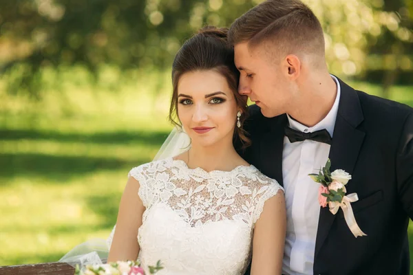 Um jovem casal de roupas de casamento está sentado em um banco em um parque. Tempo ensolarado — Fotografia de Stock