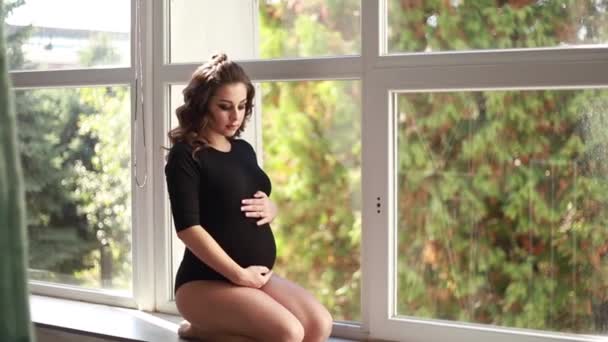 Mãe grávida senta-se na frente de uma grande janela. Modelo vestido de macacão preto. Ela pôs a mão na barriga. Futura mãe feliz — Vídeo de Stock